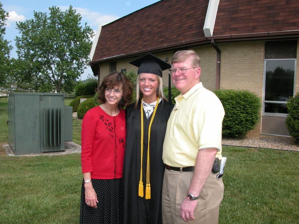 A graduate in cap and gown stands between an older man in a yellow shirt and a woman in a red cardigan in front of a building and grassy area.